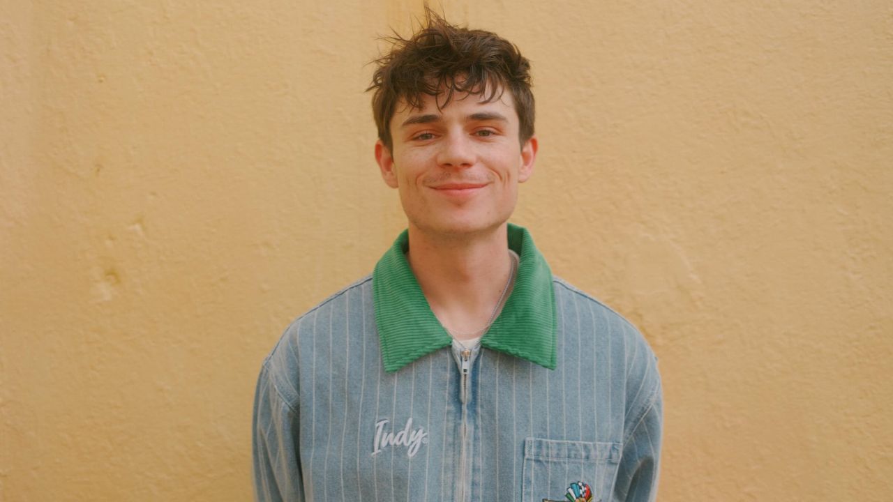 Ashby Gentry showcases a sharp retro-streetwear look with a pinstriped denim zip-up jacket accented by a bold green corduroy collar and a minimalist silver necklace.