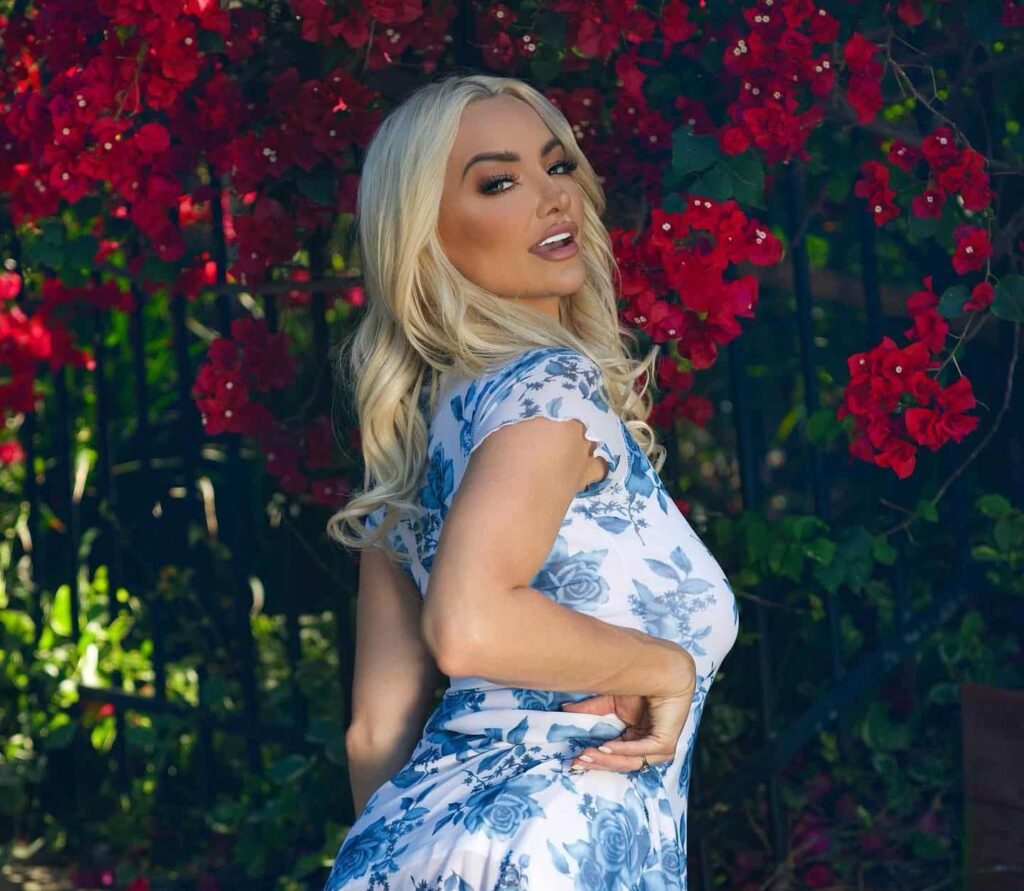 Striking a playful pose against a backdrop of vibrant red bougainvillea, Lindsey Pelas showcases timeless summer elegance in a fitted white dress adorned with a delicate blue floral pattern.