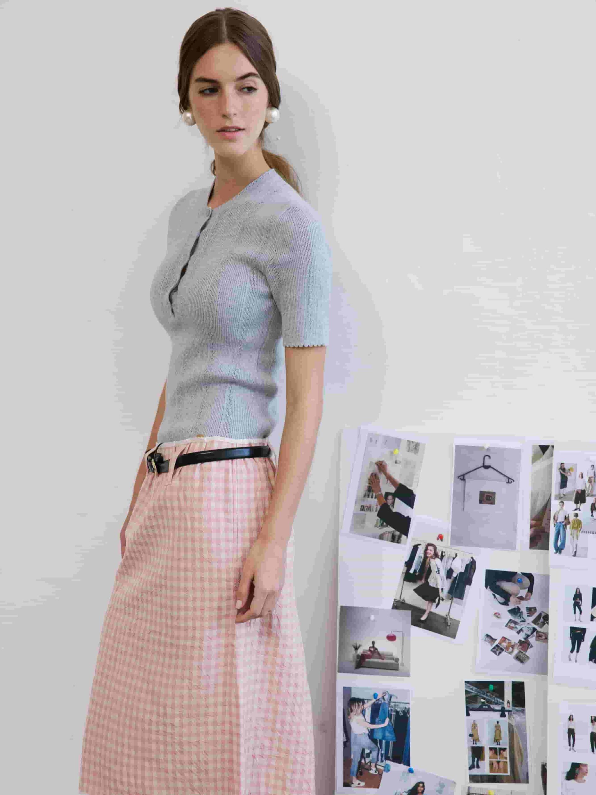 Exuding vintage-inspired charm beside a fashion mood board, Emily Feld styles a cozy short-sleeve knit top with a pastel pink gingham midi skirt and elegant statement pearl earrings.