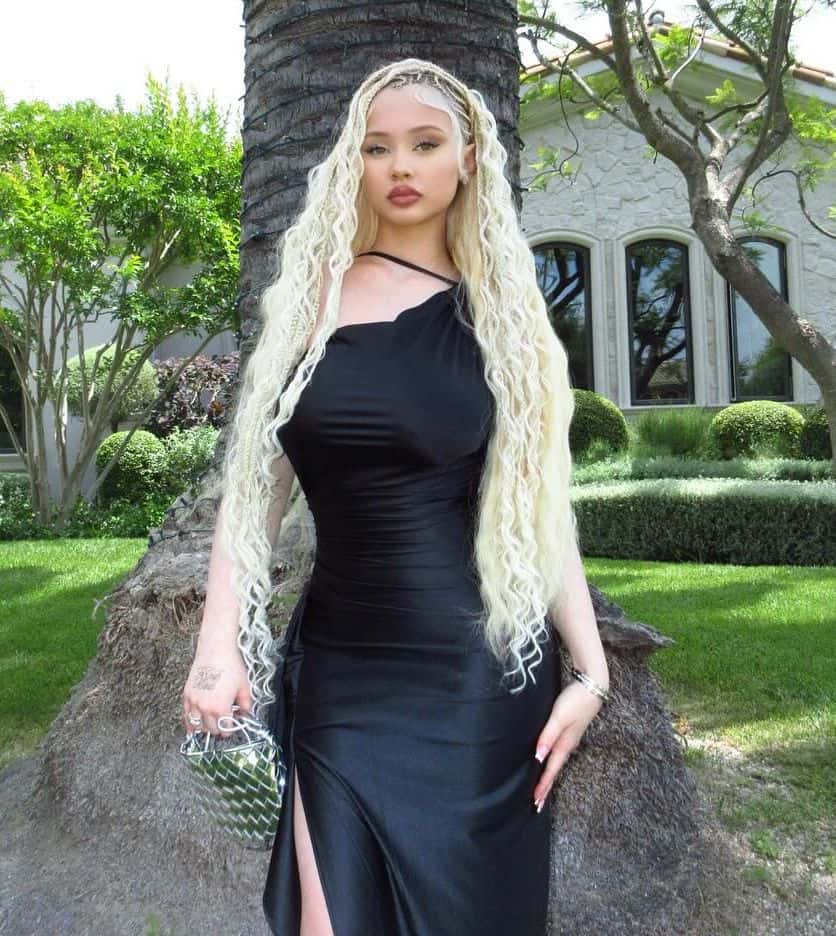 Alabama Barker channels high-fashion elegance in a sleek one-shoulder black bodycon dress paired with long textured blonde locks and a statement metallic handbag.