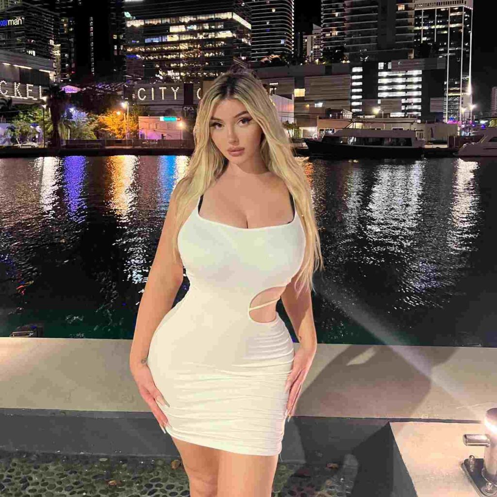 Embodying modern evening elegance, Carli Nicki wears a sophisticated white ruched mini dress with chic waist-cutout detailing against a vibrant waterfront skyline.