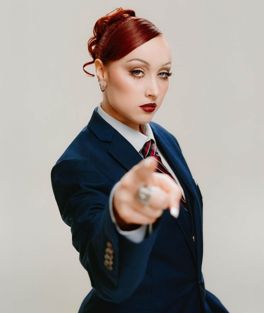 Chappell Roan commands attention in a sharply tailored navy blue power suit and striped necktie, elevated by a sculpted retro updo and striking high-glam makeup.
