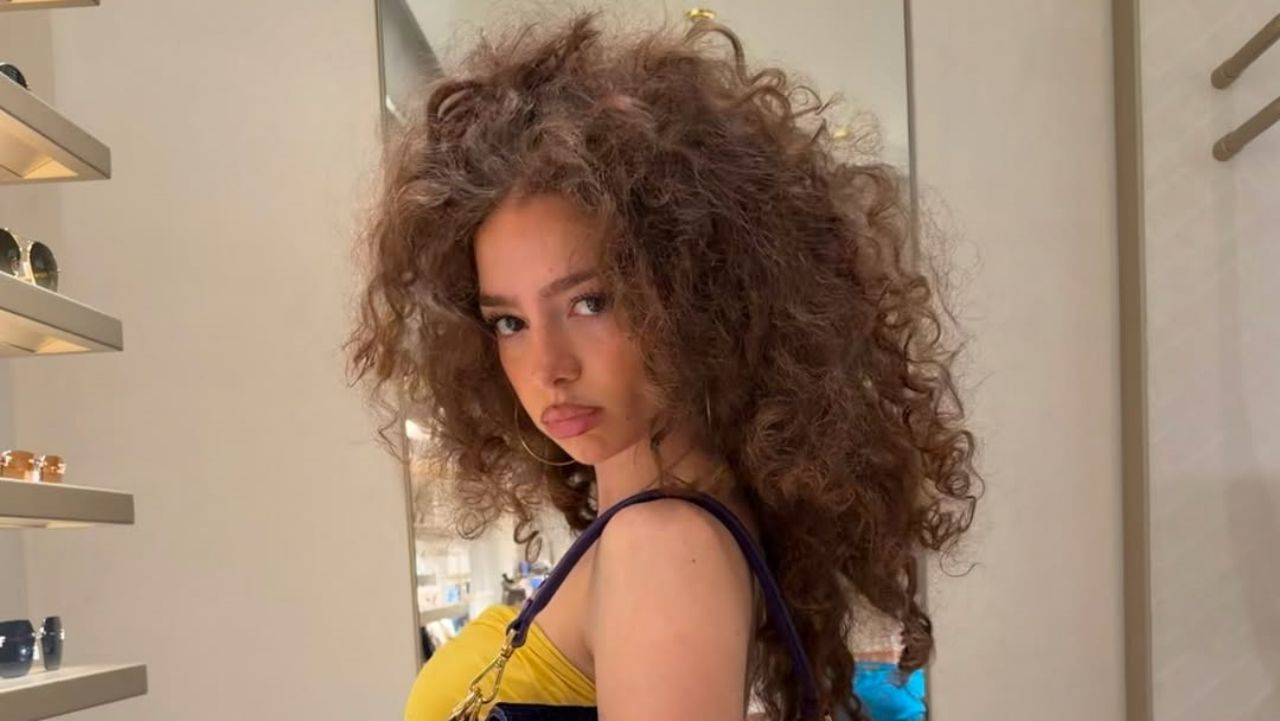 Demitra Kalogeras proves that natural hair volume is the ultimate fashion statement, perfectly framed by large gold hoops and a vibrant yellow top.