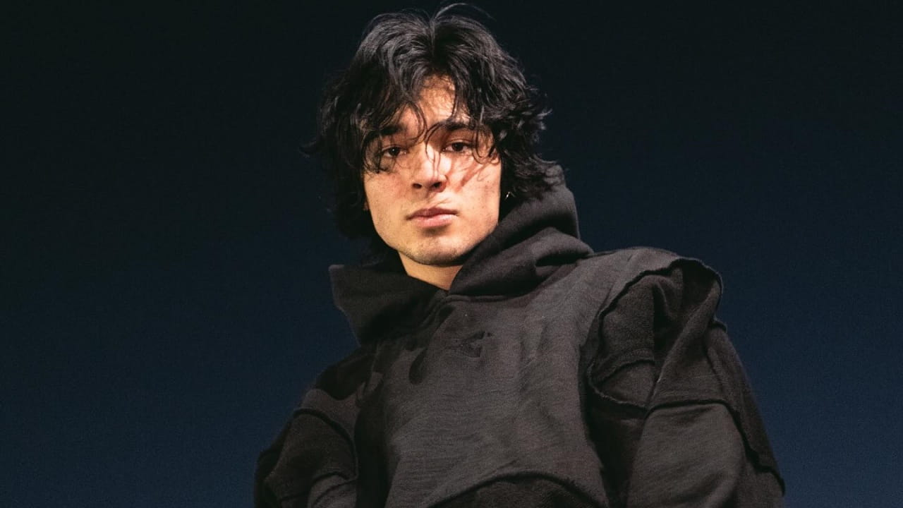 Isaac Arellanes embraces a moody streetwear aesthetic, sporting a textured black hoodie with deconstructed seam detailing and effortless tousled hair.