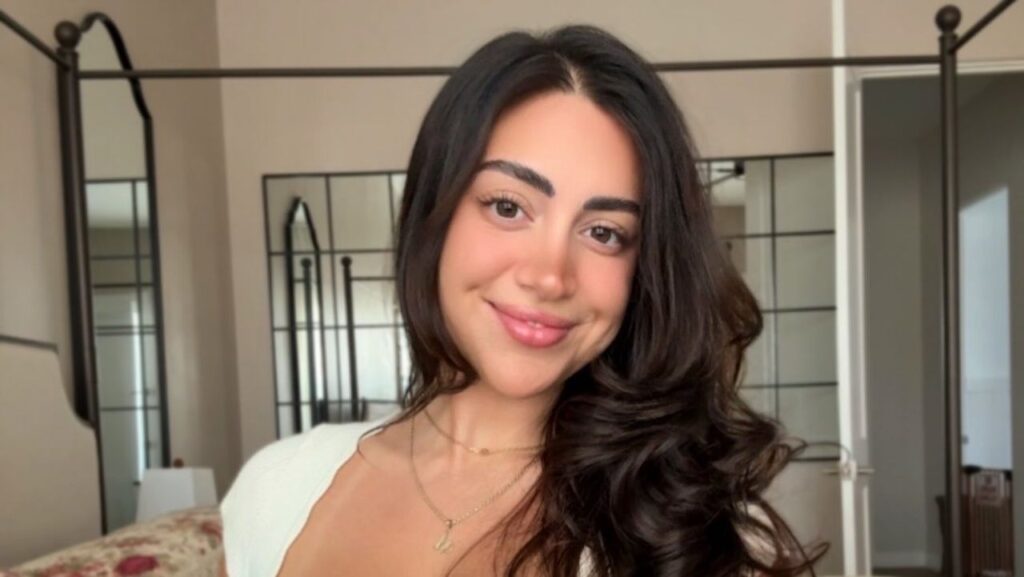 Embracing a fresh and radiant aesthetic, Mia Monroe pairs voluminous side-swept brunette waves with a clean off-white knit top and a delicate personalized initial pendant.