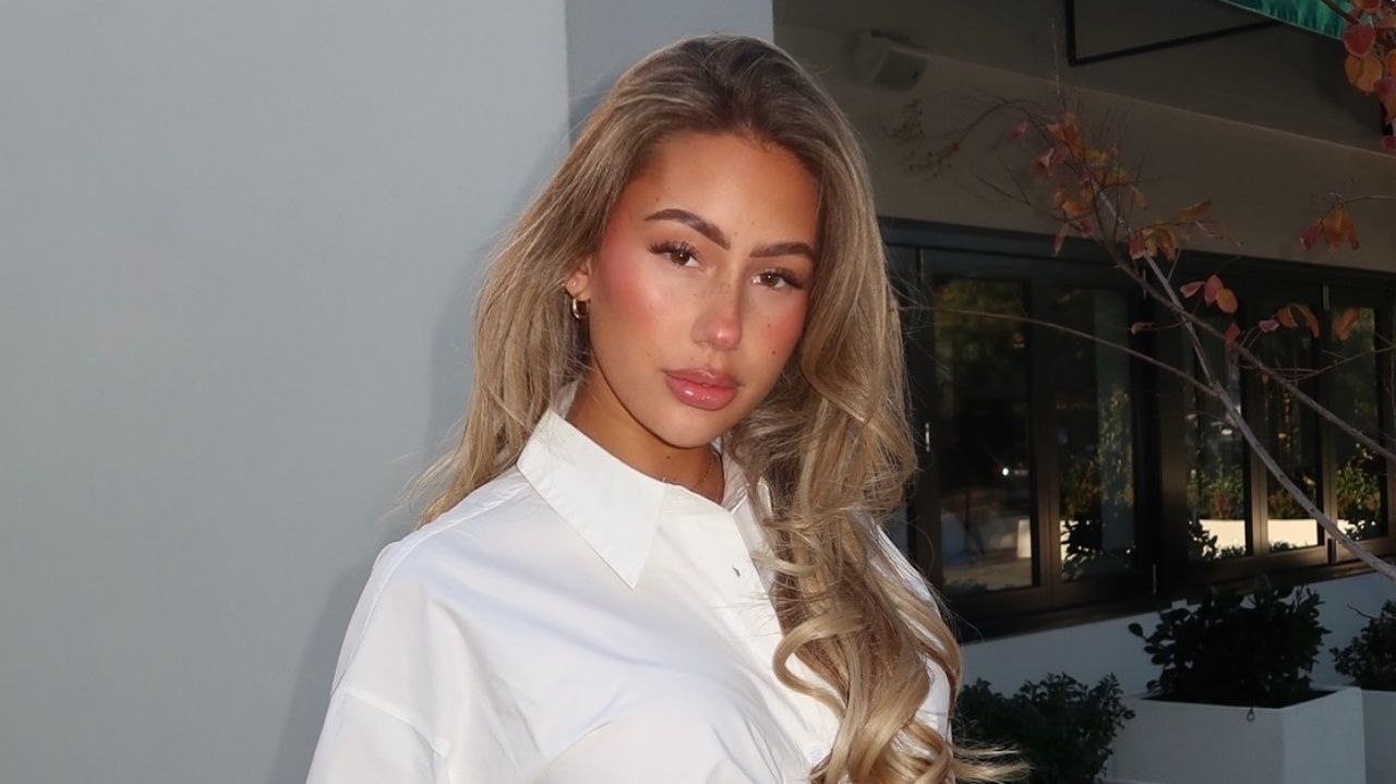 Paige Cantone radiates effortless sophistication in a crisp white button-down shirt styled with delicate gold hoops and a luminous soft-glam blowout.