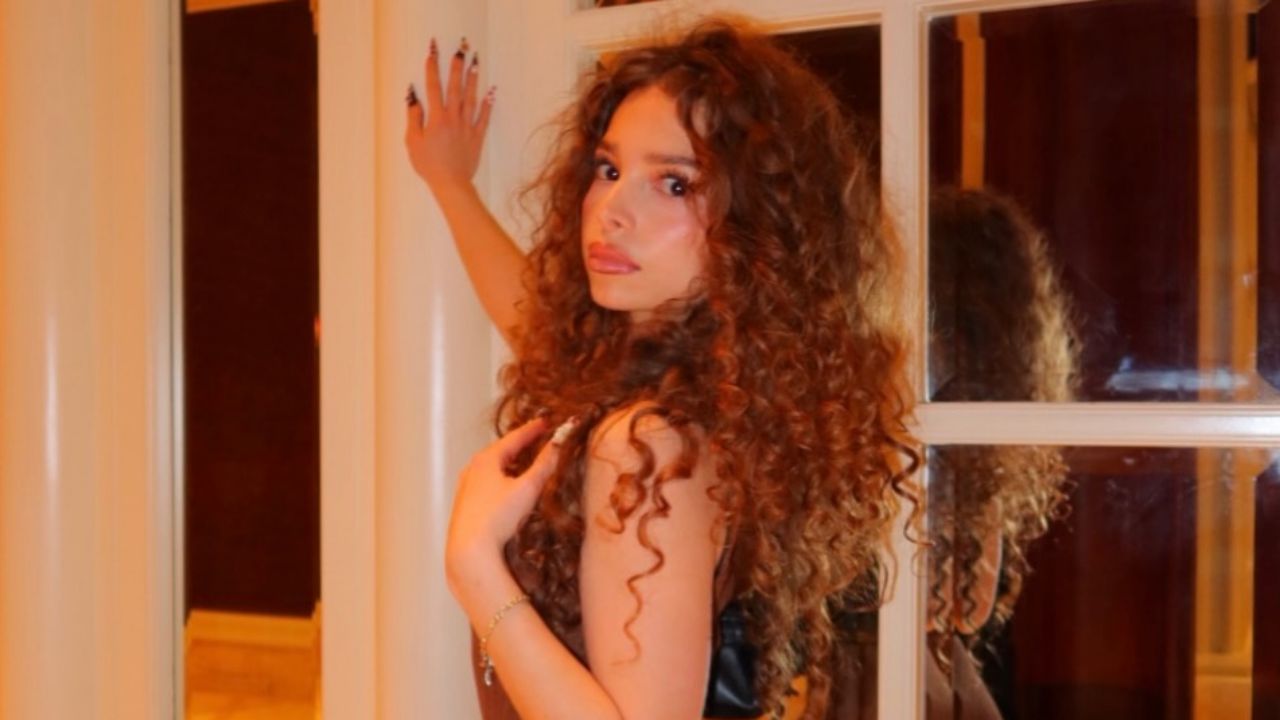 Sunday Kalogeras exudes warm elegance with her voluminous cascade of bouncy curls, glowing makeup, and delicate gold jewelry.