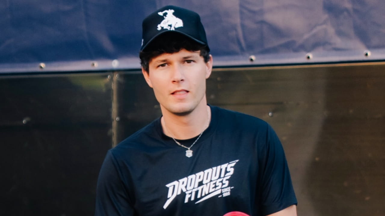 Zach Justice rocks an effortless athletic streetwear vibe, styling a classic black branded fitness tee with a structured graphic trucker cap and a delicate silver pendant necklace.