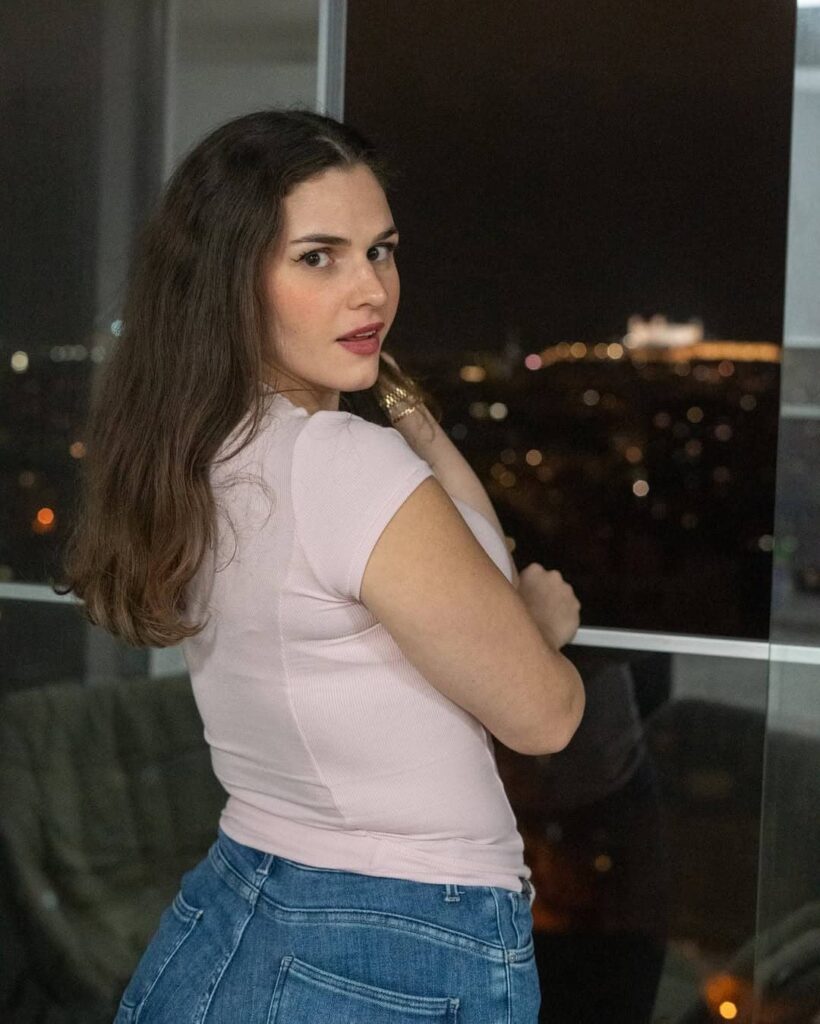 Emma Aven embraces effortless everyday chic, pairing a soft pastel pink ribbed tee with classic blue denim against a glowing city backdrop.