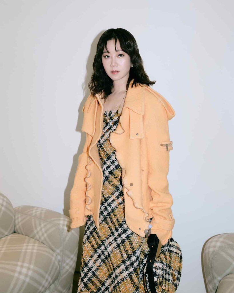 Gong Hyo Jin makes a bold fashion statement with a vibrant peach knitted jacket layered over a multi-colored plaid tweed dress, showcasing a unique and textured street-style look.