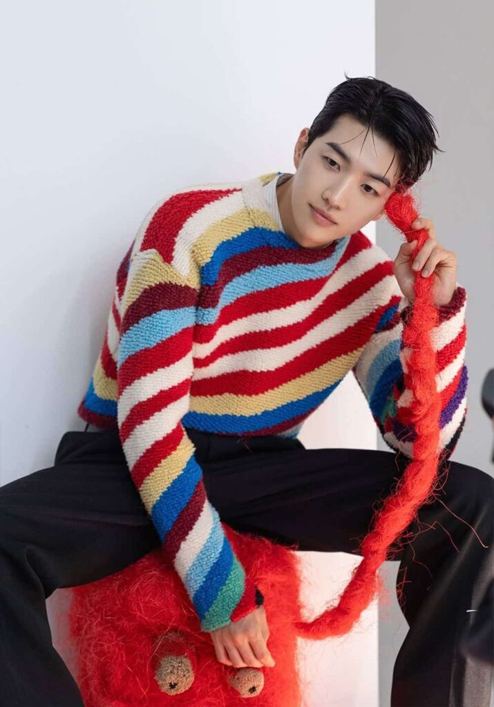 Kang Hoon embraces high-fashion editorial style with a vibrant, diagonal-striped textured knit sweater paired seamlessly against classic tailored black trousers.