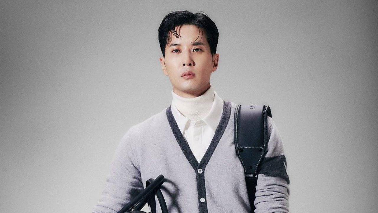 A sophisticated layered menswear ensemble, Kim Ji Suk is pictured in a grey v-neck cardigan over a white button-down polo and turtleneck, complete with a black leather messenger bag strap