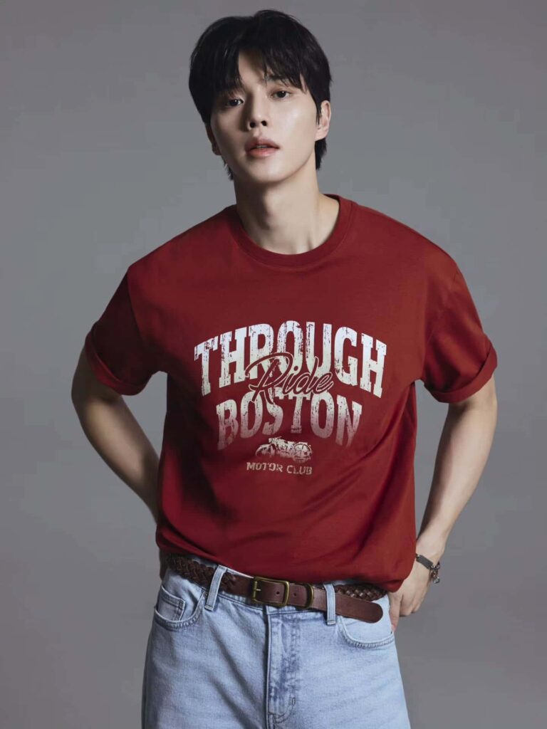 Channeling effortless retro streetwear, Song Kang pairs a vintage-inspired rust red graphic t-shirt featuring rolled-up sleeves with classic light-wash denim and a braided leather belt.